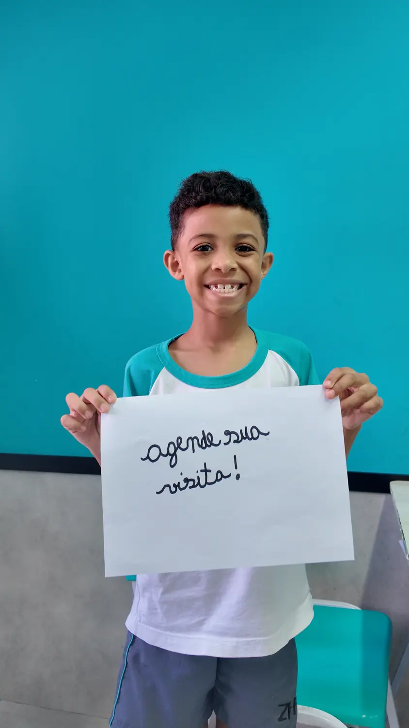 Aluno do Fundamental I sorrindo e segurando uma folha escrita à mão: agende sua visita!