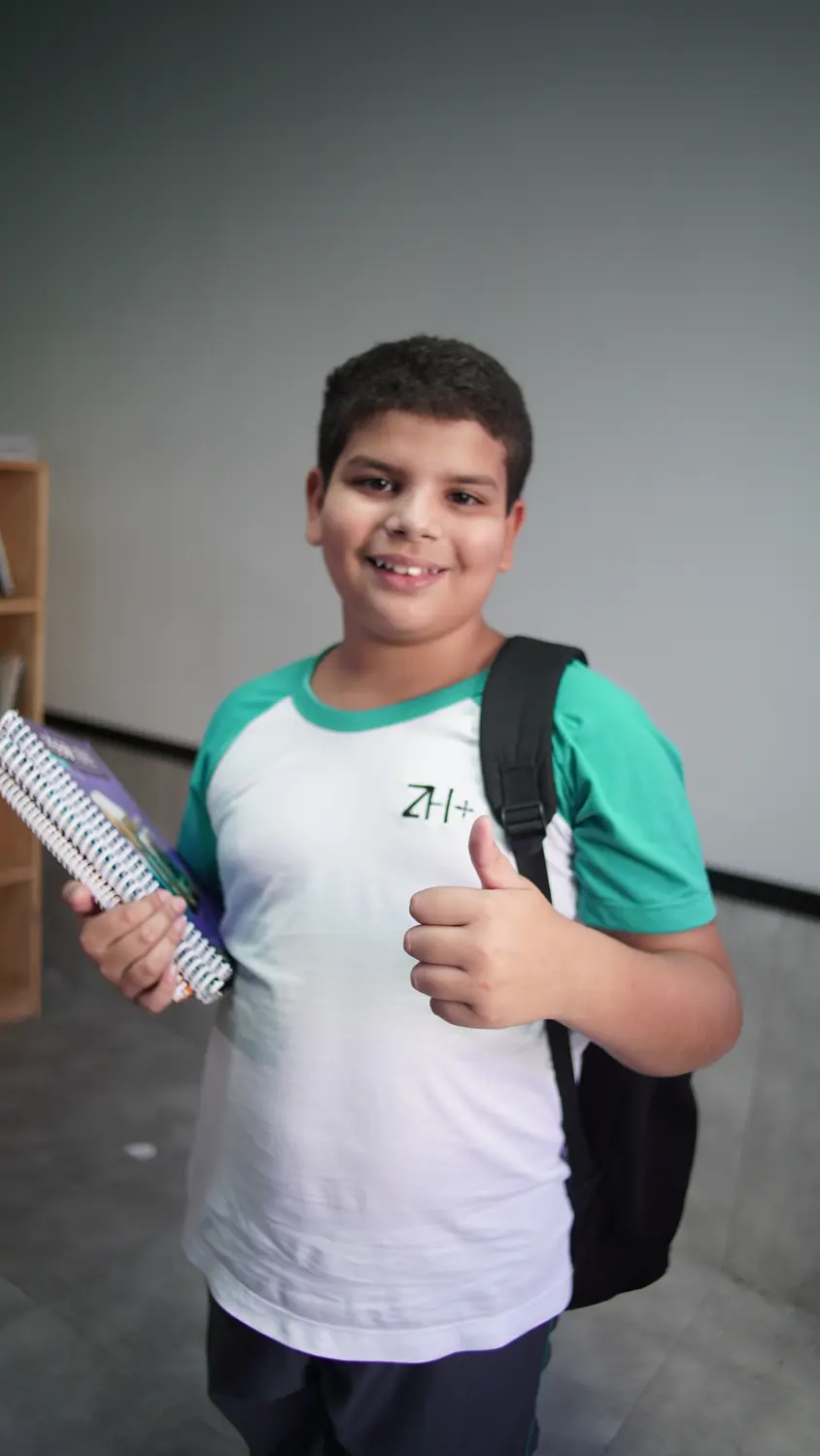 Aluno do Fundamental I do ZH+ de mochila, com o caderno na mão, fazendo sinal de positivo