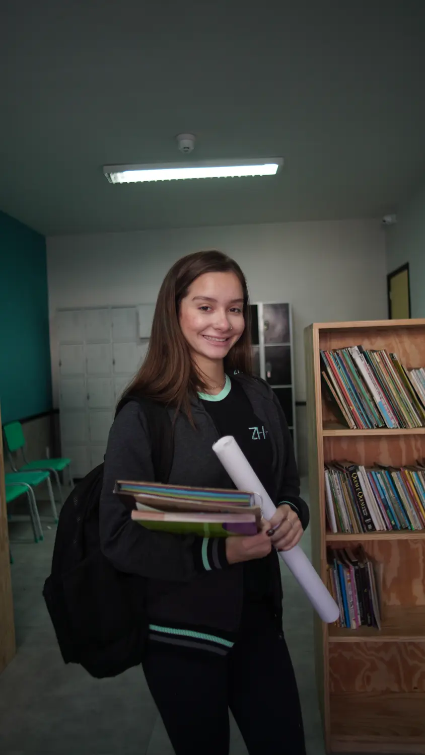 Aluna do Ensino Médio com livros, ao lado da estante da biblioteca