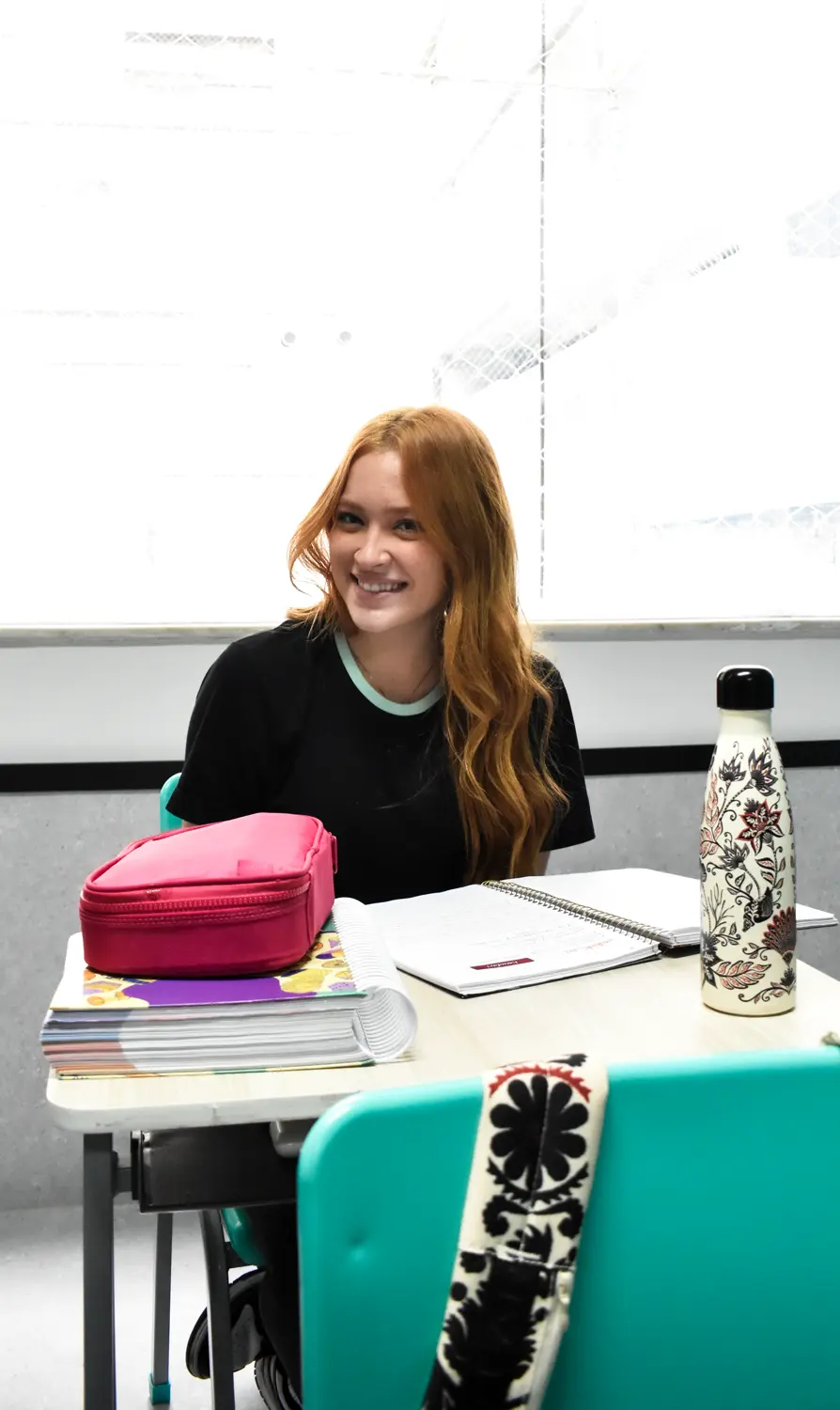 Aluna do Ensino Médio sorrindo na carteira, com cadernos e livros sobre a mesa