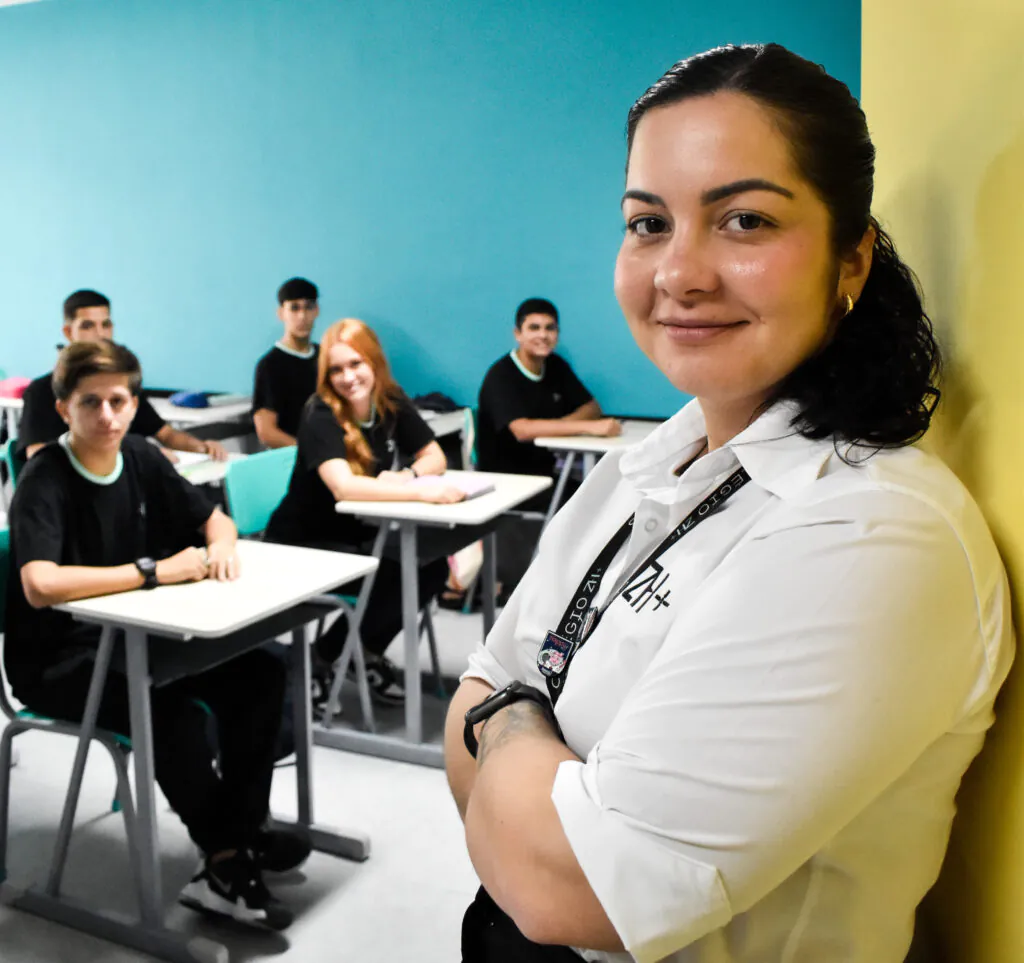 Professora sorrindo em sala, com a turma ao fundo