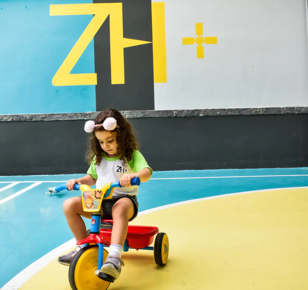 Aluna da Educação Infantil pedalando um triciclo na quadra, em frente ao mural com a marca ZH+